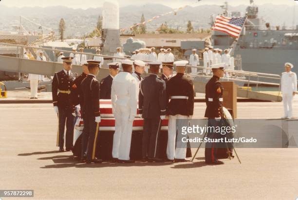 Color photograph, showing servicemen, including United States Marine Corps members, surrounding a US flag-covered coffin, resting on a dock, with...