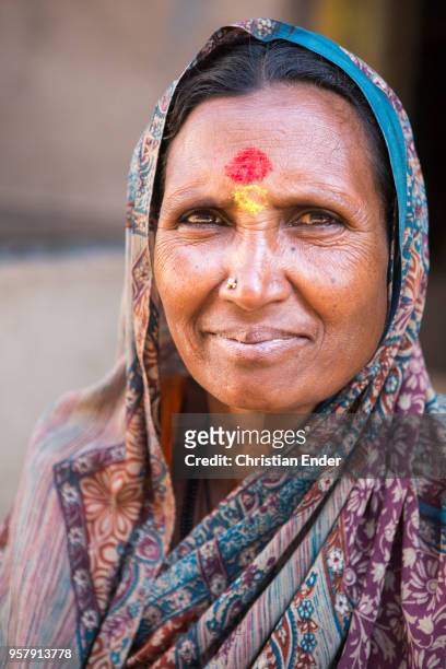 An elderly Indian woman squats in front of a house. She wears a traditional colorful hairscarf. As Hindu woman she is signed with a bindi, what...