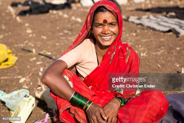 Portrait of a smiling indian woman with a traditional red scarf. She is sitting outside at a campfire site of an dry field. As Hindu woman she is...