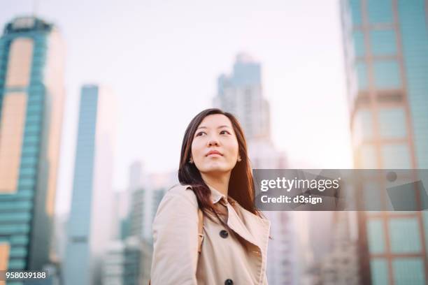 confident young asian woman looking up to sky with smile on a fresh bright morning against city skyline - smile asia woman stock-fotos und bilder