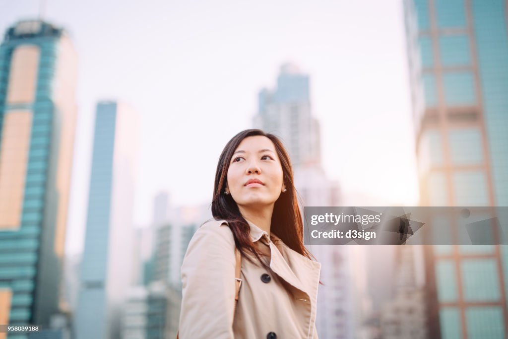 Confident young Asian woman looking up to sky with smile on a fresh bright morning against city skyline