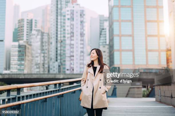 beautiful young woman walking along urban city street, against highrise modern buildings - asian woman smiling sunrise ストックフォトと画像