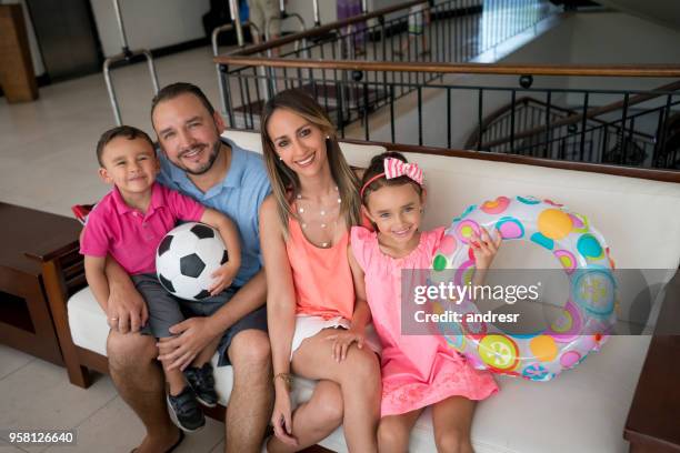 porträt der lateinamerikanischen familie sitzen auf der couch in einem hotel alle blick auf die kamera zu lächeln, während kinder einen fußball und einen aufblasbaren ring festhalten - mother of all balls stock-fotos und bilder