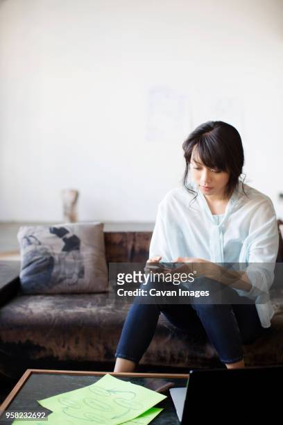 businesswoman text messaging while sitting on couch in creative office - mobile phone edit stock-fotos und bilder