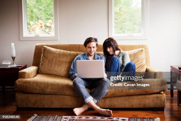 man using laptop computer while sitting with girlfriend on sofa at home - wohnzimmer frontal stock-fotos und bilder