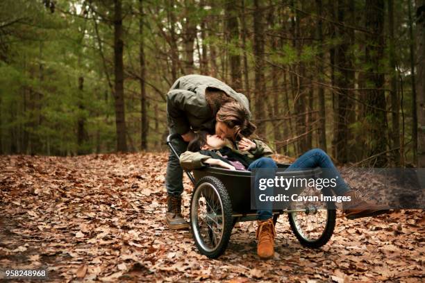 man kissing woman sitting on wheelbarrow in forest - kruiwagen stockfoto's en -beelden