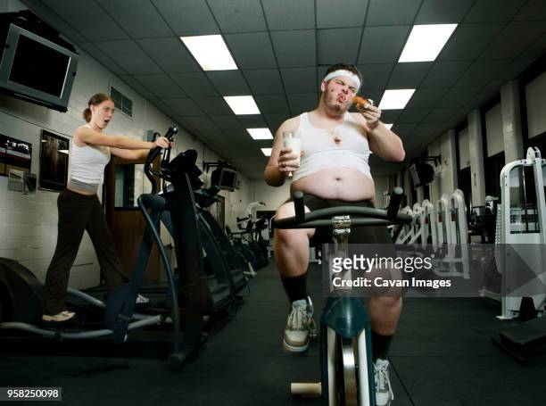 shocked woman looking at fat man eating donut while exercising in gym - fat woman funny stockfoto's en -beelden