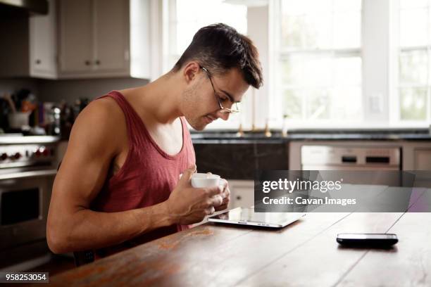 man with coffee cup looking at tablet computer while sitting by table in kitchen - man glasses tablet in kitchen stock pictures, royalty-free photos & images