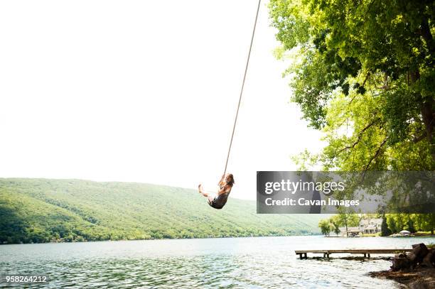 woman swinging on rope over lake against clear sky - rope swing fotografías e imágenes de stock