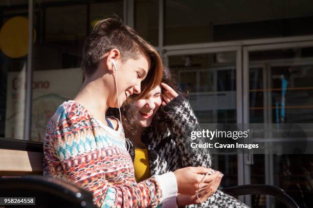 happy friends using mobile phone while sitting on bench - corte de pelo con media cabeza rapada fotografías e imágenes de stock