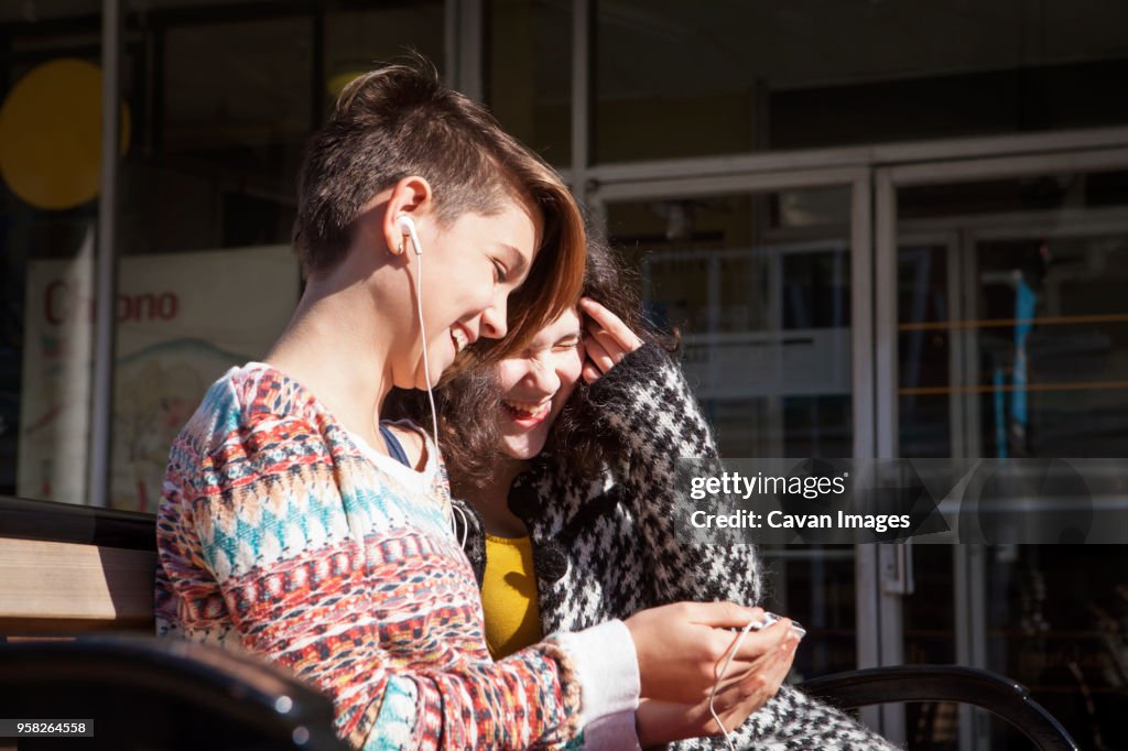 Happy friends using mobile phone while sitting on bench