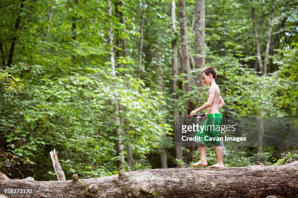 side view of boy with fishing rod walking on fallen tree trunk in forest - shorts down stock pictures, royalty-free photos & images