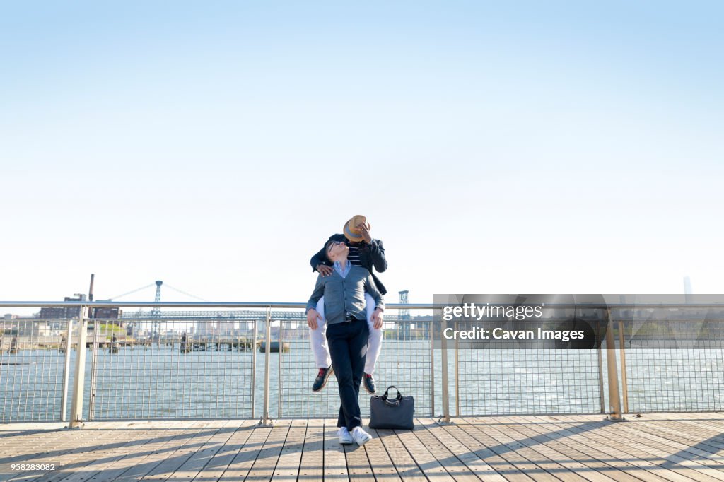 Man sitting on railing while looking at boyfriend by river against clear sky