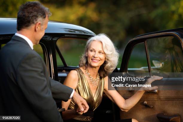 happy senior woman holding mans hand while disembarking from vintage car - holding hands in car stock pictures, royalty-free photos & images