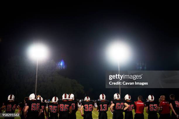 rear view of american football players standing on illuminated field against sky - safety american football player imagens e fotografias de stock