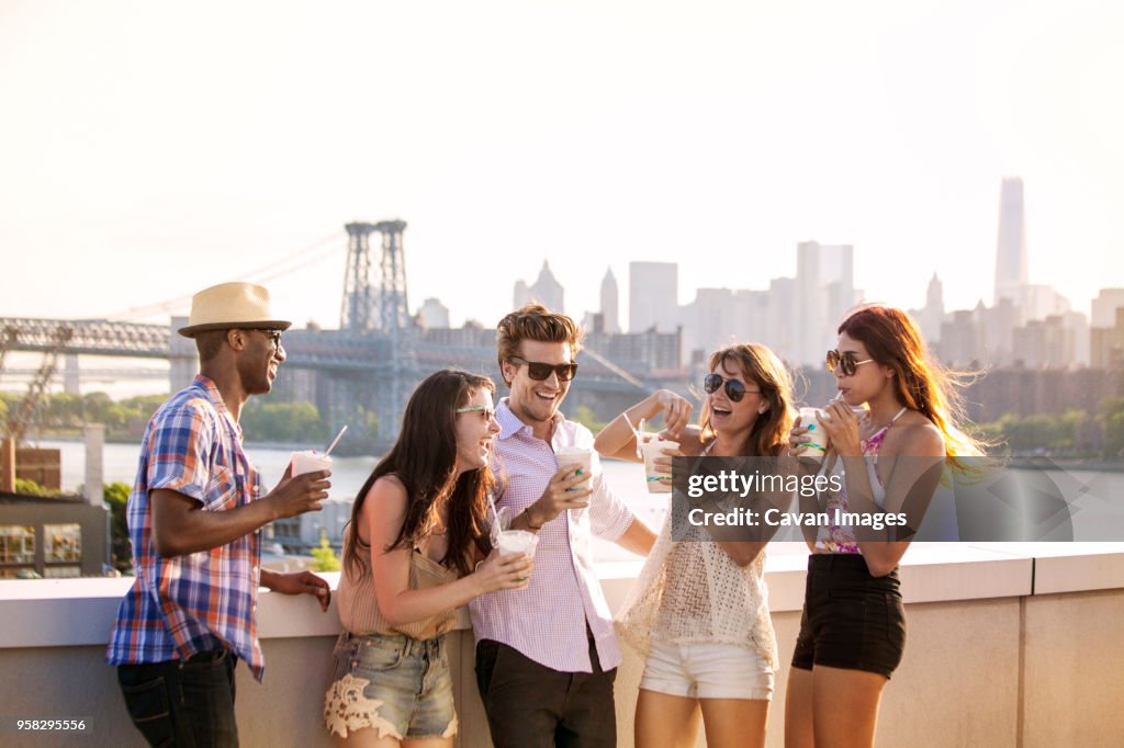 Happy friends drinking iced coffee while standing on building terrace against Williamsburg Bridge