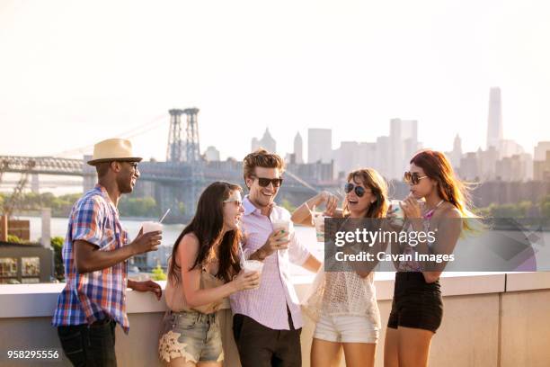 happy friends drinking iced coffee while standing on building terrace against williamsburg bridge - williamsburg new york city bildbanksfoton och bilder