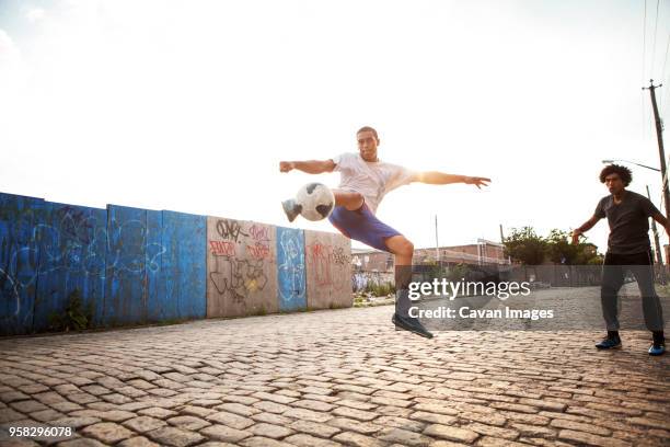 man kicking soccer ball while friend looking at street - street football stock pictures, royalty-free photos & images