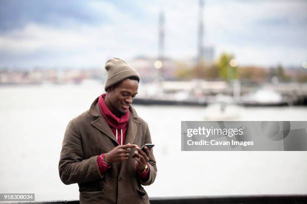 smiling man using mobile phone while standing by railing against sea - オーバーコート ストックフォトと画像