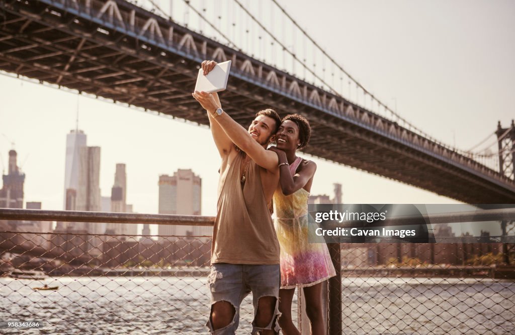 Multi-ethnic couple taking selfie through tablet computer against Manhattan Bridge and city skyline