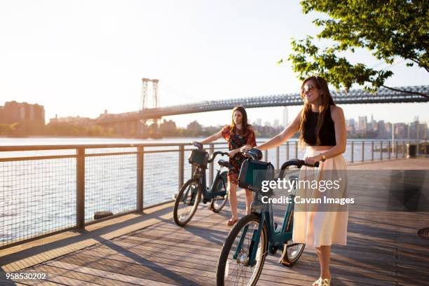 friends with bicycle walking on promenade against williamsburg bridge during sunset - williamsburg new york city stock pictures, royalty-free photos & images