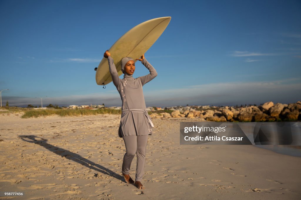 Black muslim girl wearing hijab and walking to the ocean with a surfboard