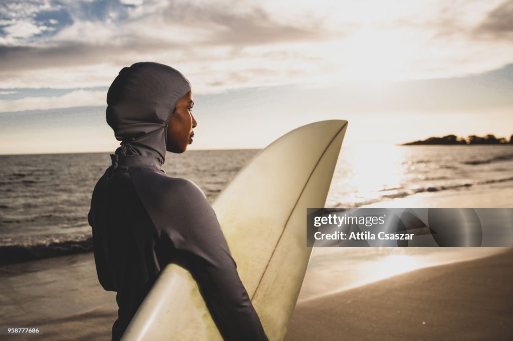 Black muslim girl wearing hijab and looking at distance while holding a surfboard