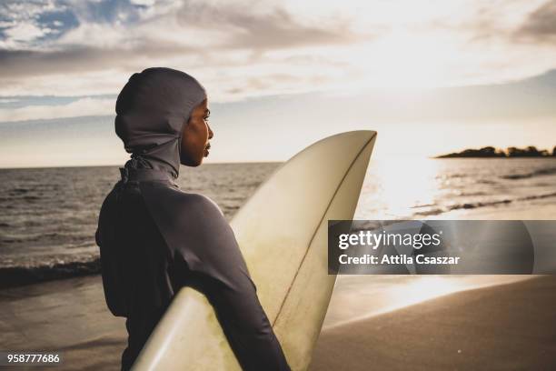 black muslim girl wearing hijab and looking at distance while holding a surfboard - traditional clothing stock pictures, royalty-free photos & images