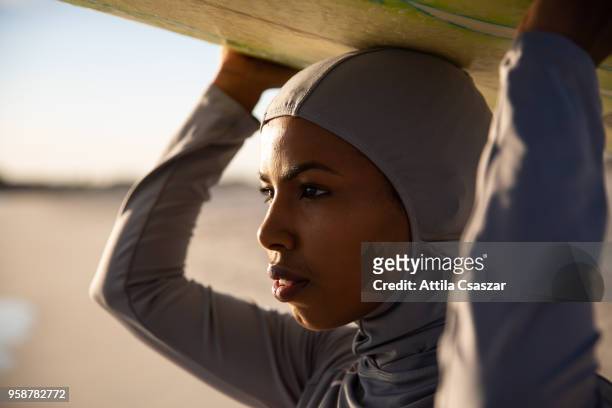 Black muslim girl wearing hijab and looking at distance while holding a surfboard