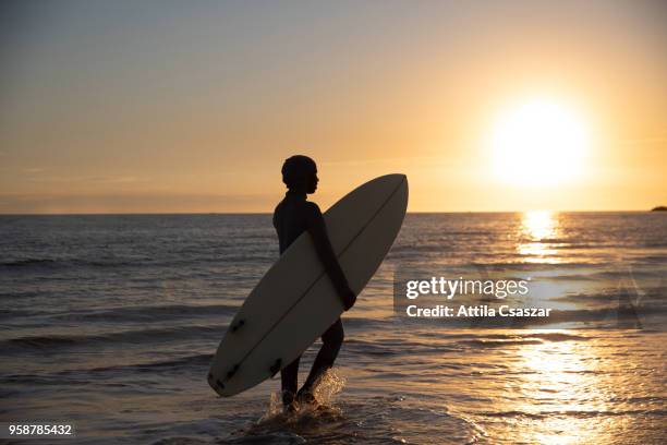 Black muslim girl wearing hijab and walking in the ocean while holding a surfboard