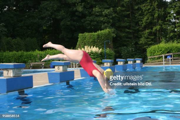 Junge Frau im Badeanzug mit Badekappe und Schwimmbrille springt von einem Startblock im Poseidon-Freibad