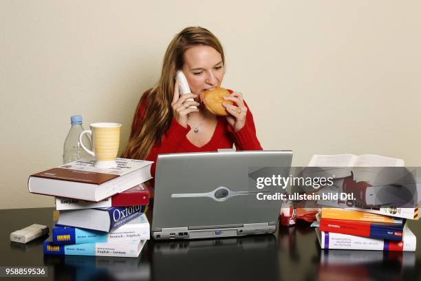 Gestresste Studentin isst beim Lernen zwischen Buechern und einem Laptop. -