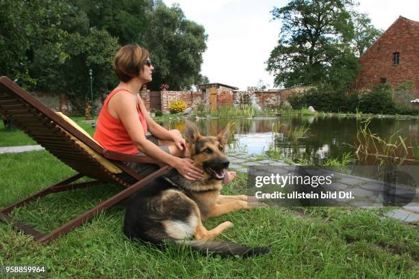 Germany - Brandenburg - Altlewin: woman with a dog is relaxing in a canvas chair at a carp pond in the garden of a farm -