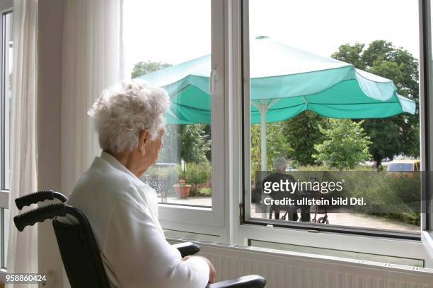 Altenheim, Altenpflege, Rentnerin im Altersheim sitzt im Rollstuhl am Fenster und schaut in den Garten -