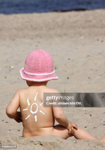 Ein Kleinkind mit Sonnencreme auf dem Ruecken sitzt am Strand