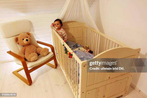 Ein Kind steht in seinem Kinderbett unterm Himmel, daneben ein Schwingsessel mit einem Teddybaer