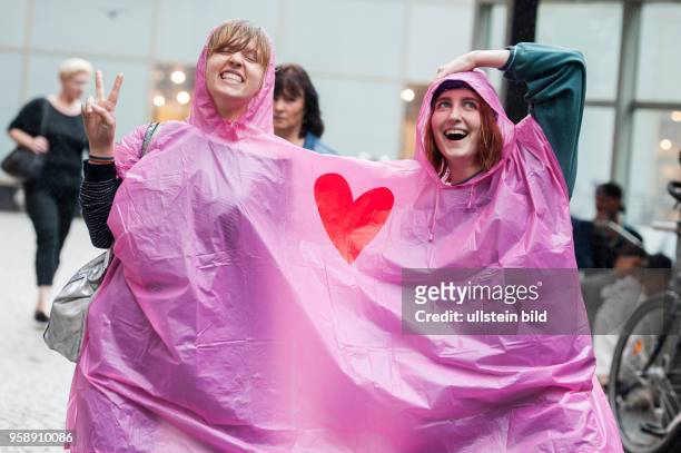 Zwei junge Frauen trotzen gut gelaunt dem Berliner Dauerregen und teilen sich ein gemeinsames Regencape