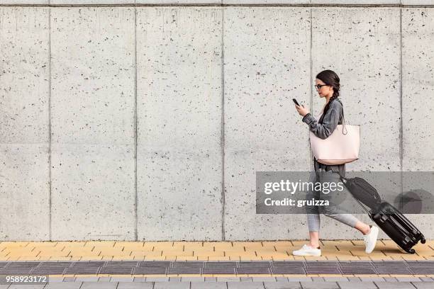 young woman using her smart phone walking beside the concrete wall and pulling a small wheeled luggage with a briefcase on it - business woman suitcase stock pictures, royalty-free photos & images