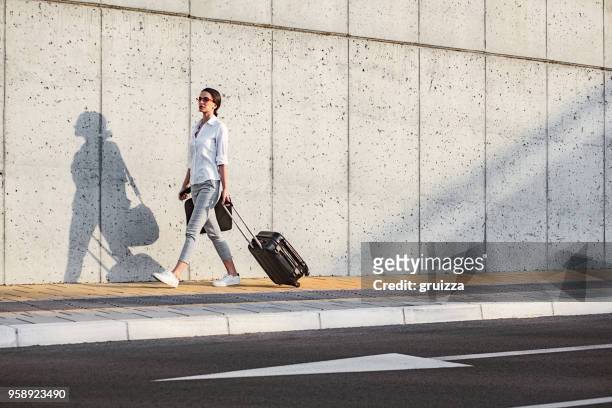 年輕女子走在人行道旁邊的混凝土牆和拉一個小輪式行李與公事包上 - wheeled luggage 個照片及圖片檔