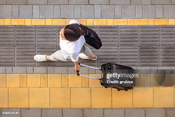 high angle view of a young woman walking on a sidewalk and pulling a small wheeled luggage with a briefcase on it - passenger stock pictures, royalty-free photos & images