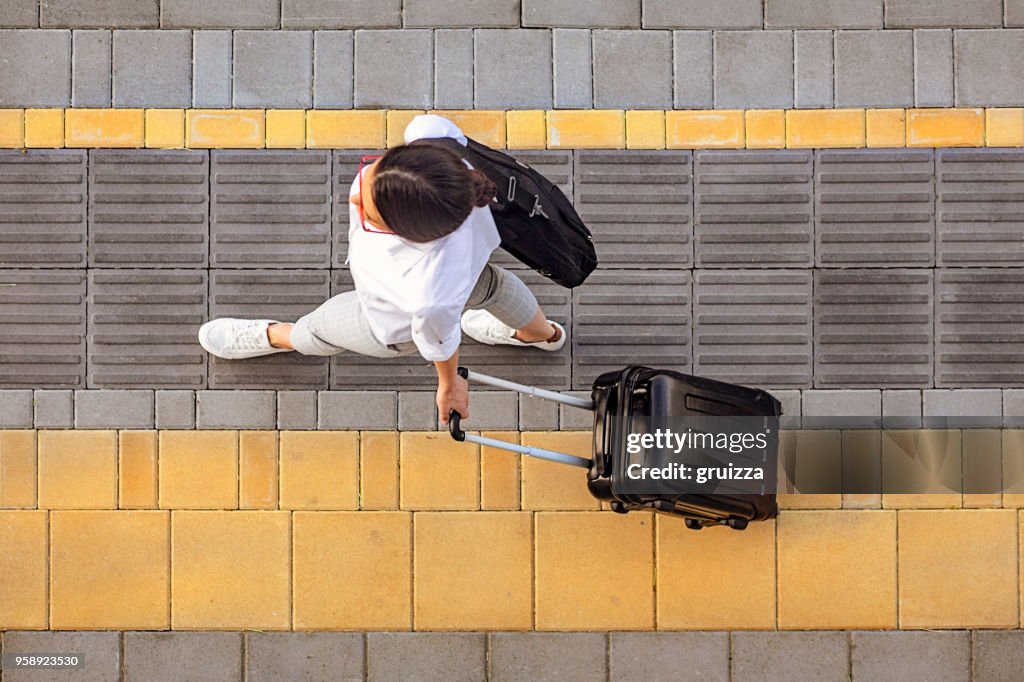 Vista de alto ângulo, de uma jovem mulher caminhando na calçada e puxando uma pequena bagagem com rodas com uma pasta nele