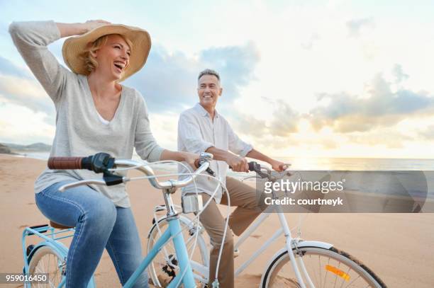 mature couple cycling on the beach at sunset or sunrise. - happy couple stock pictures, royalty-free photos & images