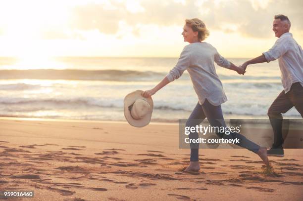 mature couple running and playing on the beach at sunset or sunrise. - couple running on beach stock pictures, royalty-free photos & images