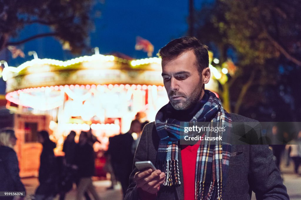 Handsome man using smart phone in front of illuminated carousel