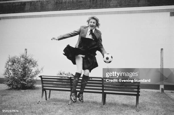 Scottish soccer player Denis Law jumping over a bench while wearing a traditional Scottish kilt and holding a soccer ball, UK, 24th May 1978.