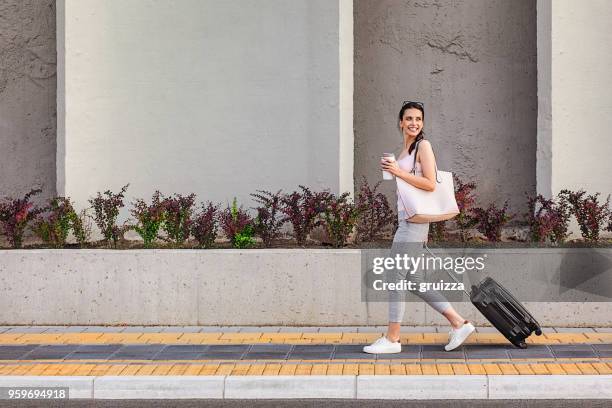 young woman walking on a sidewalk beside the concrete wall and pulling a small wheeled luggage - wheeled luggage stock pictures, royalty-free photos & images