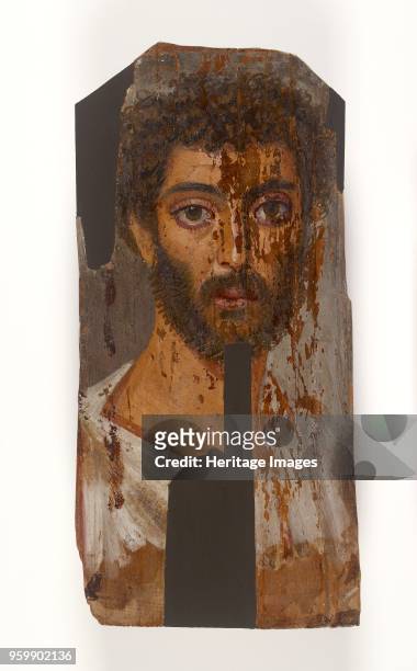 Mummy portrait, 2nd Century. Portrait painted in encaustic on thin panel, showing young man, thick curly hair and beard, white drapery ith red...