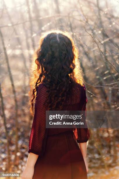 back view of young woman with long wavy red hair wearing 3/4-sleeve burgundy dress, sunlit in forest - anna gorin stock-fotos und bilder