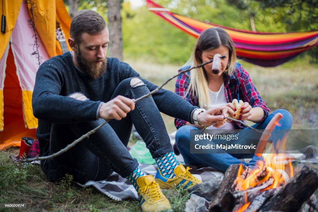 Beautiful couple, sitting, camping around the campfire. On the background with a tent The man flicks the fire with a stick.