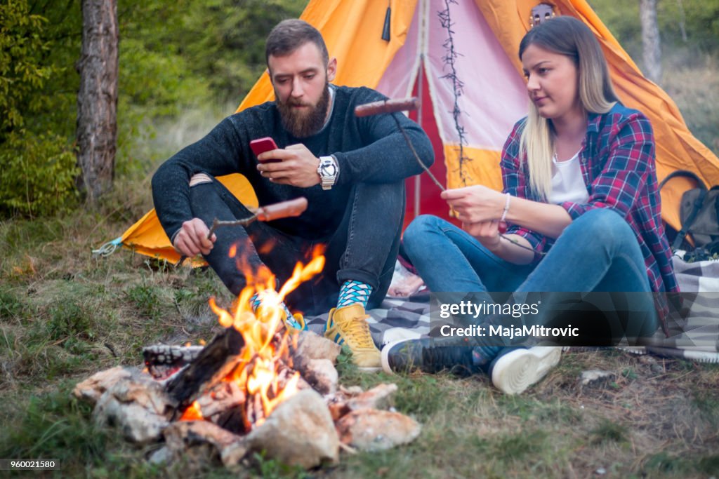 Beautiful couple, sitting, camping around the campfire. On the background with a tent The man flicks the fire with a stick.